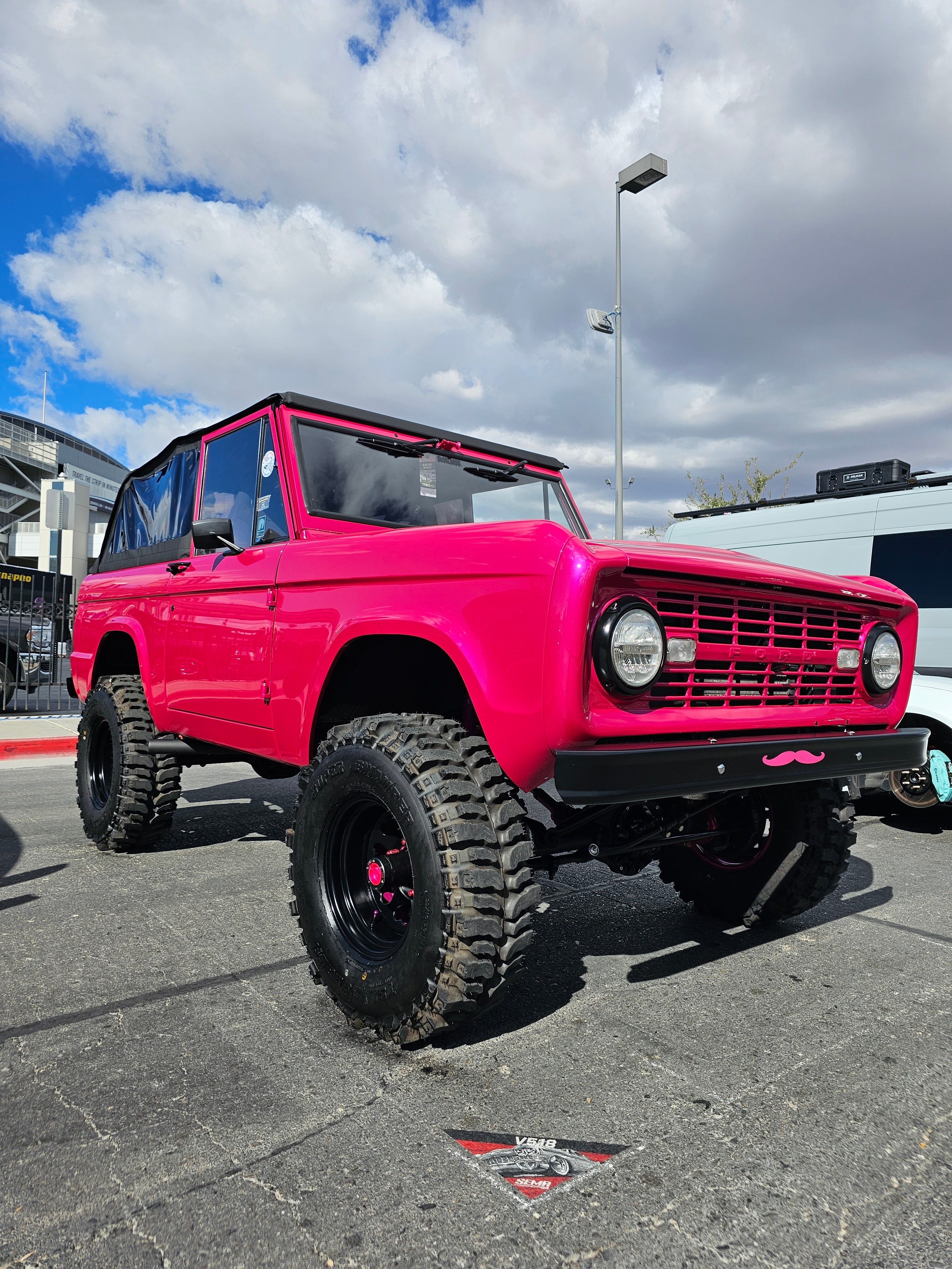 A bright pink Bronco with a pink mustache decal below the grille sits in a lot.